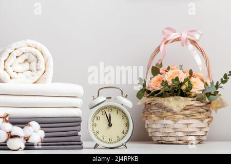 Una pila di lenzuola bianche e grigie, lenzuola e un asciugamano in spugna. Sveglia e un cestino di fiori sul tavolo. Foto Stock