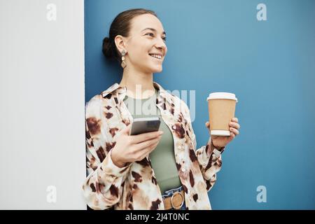 Vivace ritratto minimal di giovane donna sorridente che tiene la tazza di caffè contro il muro blu con sorriso candido, spazio copia Foto Stock