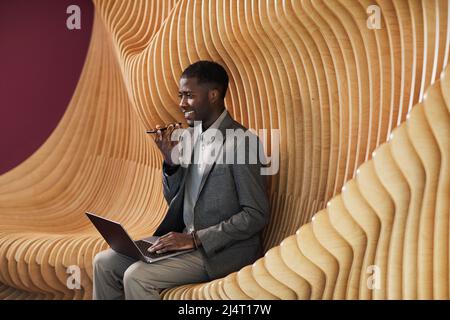 Ritratto grafico di uomo d'affari nero sorridente che registra il messaggio vocale mentre lavora nello spazio moderno dell'ufficio con le sedie del progettista, spazio di copia Foto Stock