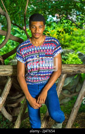 Ritratto di giovane uomo Nero. Indossando una camicia colorata modello, jeans blu, collana, un giovane ragazzo bello si trova in piedi contro una recinzione di legno, affascinante Foto Stock