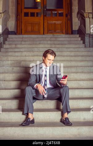 Uomo d'affari che ti aspetta. Vestirsi formalmente in abito grigio, bianco sotto la camicia, cravatta da collo a motivi geometrici, un giovane uomo d'affari è casualmente seduto sui passi o Foto Stock