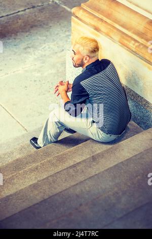 Giovane uomo che si rilassa all'esterno. Indossando una camicia a motivi neri, pantaloni grigi, scarpe in pelle, un ragazzo giovane con barba, capelli gialli, è seduto sulle scale, bac Foto Stock