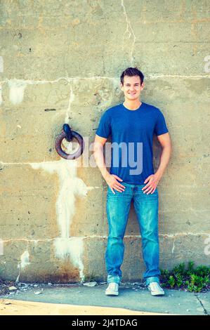 Uomo Moda casual. Indossando una T-shirt blu, jeans, sneakers bianchi, un giovane ragazzo bello si trova in piedi contro un vecchio muro di cemento con un Ri di metallo arrugginito Foto Stock