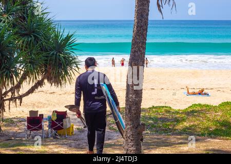 Un uomo cammina con una tavola da surf al mare lungo la costa tropicale. Viaggi e turismo, ricreazione e divertimento. Foto Stock