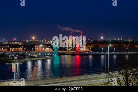 I tre vecchi porti si gru dall'isola di Szczecin di Lasztownia colorfully illuminato. Città di notte. Polonia. Dalla distanza che assomiglia ai dinosauri. P Foto Stock