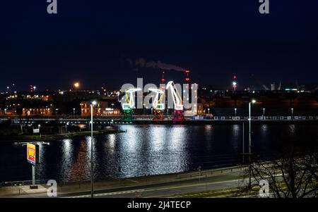 I tre vecchi porti si gru dall'isola di Szczecin di Lasztownia colorfully illuminato. Città di notte. Polonia. Dalla distanza che assomiglia ai dinosauri. P Foto Stock