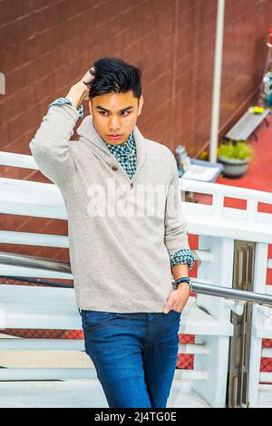 Uomo solitario. Vestirsi in maglione grigio chiaro con cardigan con colletto scialle, jeans blu, una testa graffiante a mano, una mano in tasca, un ragazzo giovane è sta Foto Stock