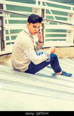 Uomo che ti aspetta. Vestirsi in maglione con shawl colletto cardigan, jeans, rotolando su polsino, un ragazzo giovane è seduto su scale da recinzione, talki Foto Stock