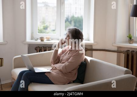 La donna più anziana si siede sul divano con il laptop pensa, fissando da parte Foto Stock