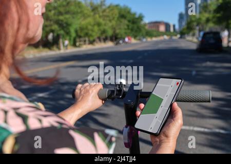 Donna matura irriconoscibile che guarda una mappa in un'app sul suo telefono per guidare il suo scooter elettrico in una città soleggiata. Concetti: Tecnologia, applicazione telefono Foto Stock