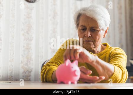 Colpo interno medio di una donna anziana dai capelli corti in un maglione giallo che mette le sue monete nella banca rosa del piggy. Concetto di risparmio. Foto di alta qualità Foto Stock