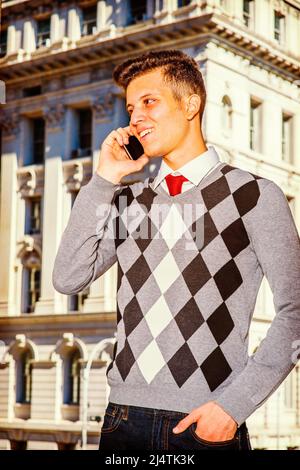 Uomo che chiama all'esterno. Vestendo in un nero, bianco, grigio, pullover a motivi geometrici, jeans, un giovane uomo d'affari bello è in piedi davanti ad un bui ufficio Foto Stock