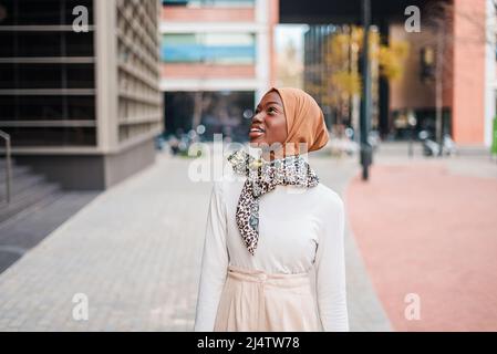 Curiosa femmina islamica afroamericana in foulard tradizionale che guarda in alto edificio nel moderno quartiere cittadino Foto Stock