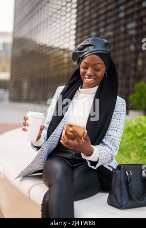 Positiva femmina afroamericana in abiti casual e sciarpa musulmana seduta con le gambe incrociate e avere caffè da asporto mentre si guarda il video sul cellulare Foto Stock