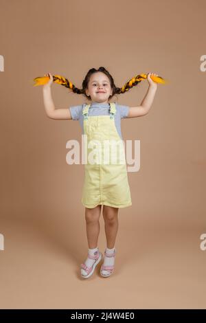 Giovane ragazza eccitato che tiene le trecce gialle del kanekalon, circa per saltare giocando guardando la macchina fotografica sorridente che indossa il jumpsuit giallo e la t-shirt grigia sul beige Foto Stock