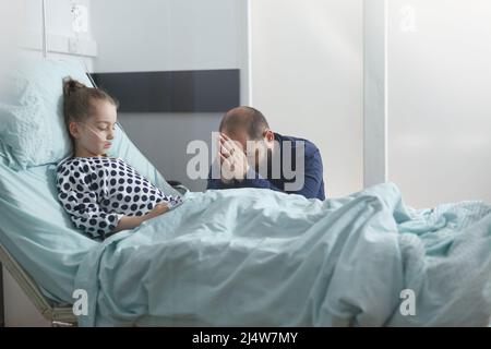 Preoccupared uneasy giovane padre che prega per ricoverato malato ragazza paziente mentre in stanza medica. Malato figlia piccola ragazza recupero da malattia mentre respira attraverso il tubo di ossigeno. Foto Stock