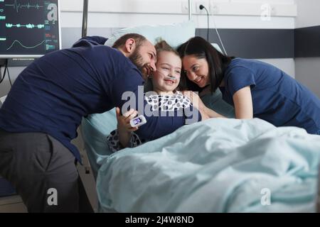 I giovani genitori felici che abbracciano malato ricoverato ricoverando la bambina mentre nella stanza pediatrica del paziente dell'ospedale. Famiglia gioiosa positiva ridendo e abbracciando nella clinica pediatrica di salute. Foto Stock