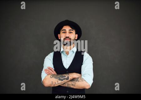 Ritratto da studio di bel maschio etnico con braccia tatuate incrociate, vestito in una camicia blu e cappello nero. Giovane uomo elegante su sfondo scuro Foto Stock