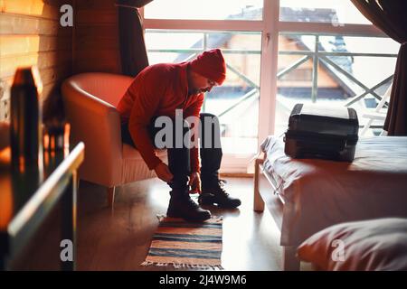 Uomo viaggiatore in un viaggio d'inverno indossando abiti rossi seduti in una camera d'albergo, legando scarpe. Con thermos sul tavolo Foto Stock