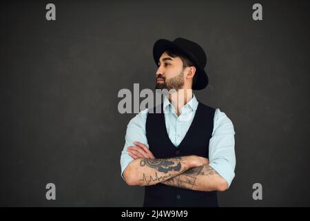 Ritratto da studio di bel maschio etnico con braccia tatuate incrociate, vestito in una camicia blu e cappello nero. Giovane uomo elegante su sfondo scuro Foto Stock