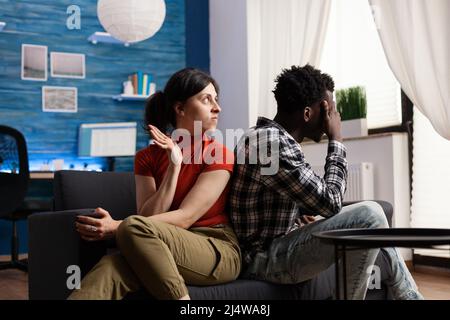 Back to back vita partner che sono arrabbiati e non parlare l'un l'altro per conflitto di relazione. Coppia interracial sconvolto che si confronta circa la cattiva situazione di matrimonio mentre a casa in soggiorno. Foto Stock