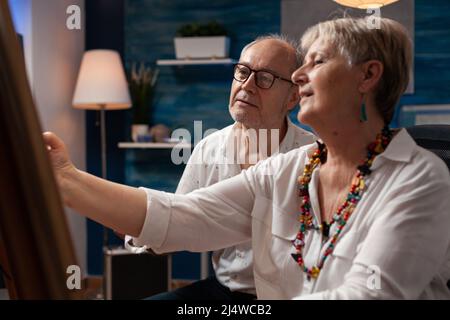 Ritratto di coppia creativa anziana disegno a matita seduta di fronte al cavalletto parlare di composizione in casa studio d'arte. Uomo più anziano che guarda la donna grigia pelata facendo schizzo di carbone. Foto Stock