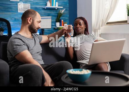 Marito attento che offre sorridente moglie incinta un bicchiere d'acqua mentre ha un portatile in grembo. Futura madre afroamericana che riceve l'acqua offerta da amorevole marito caucasico mentre si siede nel soggiorno di casa. Foto Stock