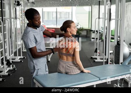 Ritratto di terapista riabilitazione mettendo cinesio nastro sulla schiena e spalla della giovane donna in clinica di fisioterapia, copia spazio Foto Stock