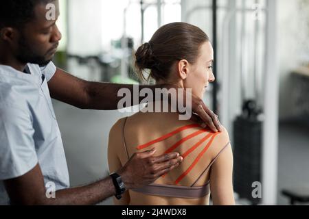 Primo piano terapista riabilitazione mettendo cinesio nastro sulla spalla della giovane donna in clinica fisioterapica, copia spazio Foto Stock