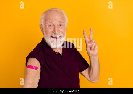 Foto di uomo allegro invecchiato mostra dita fresco v-simbolo vaccinato protezione covid isolato su sfondo di colore giallo Foto Stock