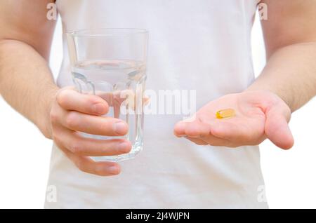 Primo piano di un uomo che indossa una maglietta bianca che tiene una capsula di olio di pesce e un bicchiere d'acqua isolato su bianco. Foto Stock