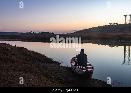 uomo seduto in tradizionale barca di legno al lago calmo con drammatico riflesso colorato cielo al mattino Foto Stock