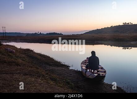 uomo seduto in tradizionale barca di legno al lago calmo con drammatico riflesso colorato cielo al mattino Foto Stock