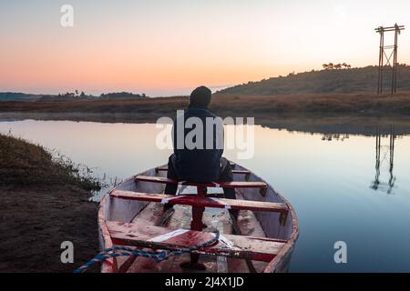 uomo seduto in tradizionale barca di legno al lago calmo con drammatico riflesso colorato cielo al mattino Foto Stock