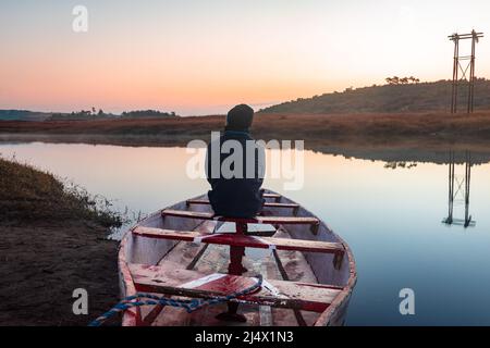 uomo seduto in tradizionale barca di legno al lago calmo con drammatico riflesso colorato cielo al mattino Foto Stock