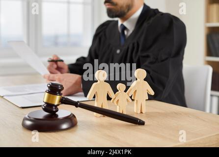 Gavel e piccole figure di famiglia sul tavolo in aula di corte, con giudice seduto sullo sfondo Foto Stock