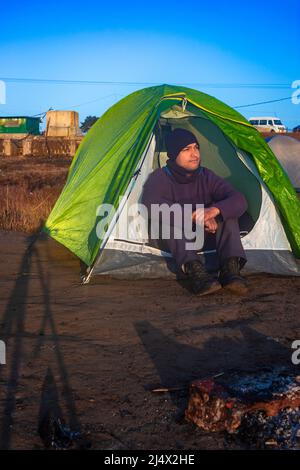 isolato giovane uomo seduto in tenda da campeggio con cielo blu luminoso al mattino da angolo piatto Foto Stock