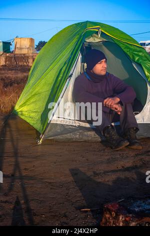 isolato giovane uomo seduto in tenda da campeggio con cielo blu luminoso al mattino da angolo piatto Foto Stock