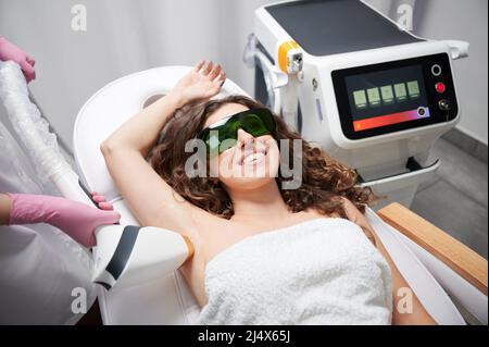 Giovane donna in occhiali di sicurezza sdraiati sul letto da giorno e sorridenti mentre estetista rimuovere i capelli da ascella femmina con dispositivo laser. Cosmetologo che esegue la procedura di rimozione dei capelli laser in salone di bellezza. Foto Stock