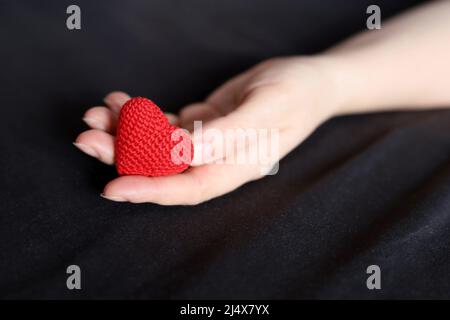 Cuore in maglia rossa nel palmo femminile di mano su un letto con biancheria da letto nera. Concetto di amore, assistenza sanitaria, maternità, donazione di sangue Foto Stock