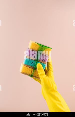 Una mano in un guanto di gomma giallo contiene spugne colorate per piatti da cui scorre il gel verde per il lavaggio delle stoviglie. Foto Stock