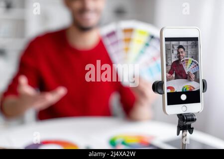 Il giovane blogger arabo sorridente con barba mostra la tavolozza dei colori e registra video sullo smartphone in salotto Foto Stock
