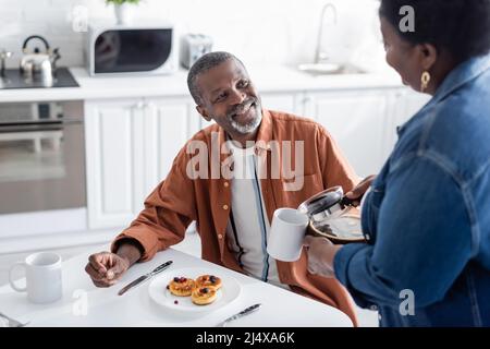 happy african american man guardando la moglie che serve caffè durante la colazione Foto Stock