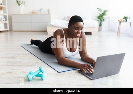 Plump giovane donna nera sdraiata su tappeto sportivo, utilizzando un computer portatile, parlando per allenare online o guardando il video di fitness a casa Foto Stock