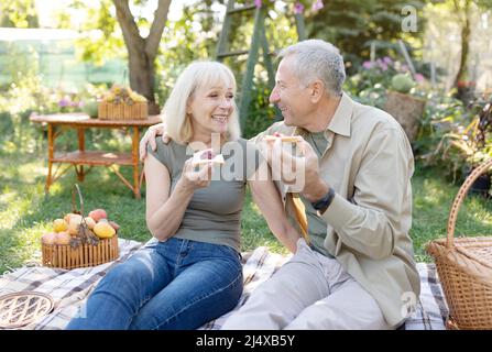 Coppia felice di età che hanno pic-nic nel loro giardino, seduta su coperta e mangiare toast con marmellata, godendo giorno di primavera Foto Stock