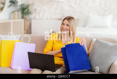 Soddisfatto giovane donna bionda europea in abiti gialli con i look per laptop nel pacchetto shopping Foto Stock
