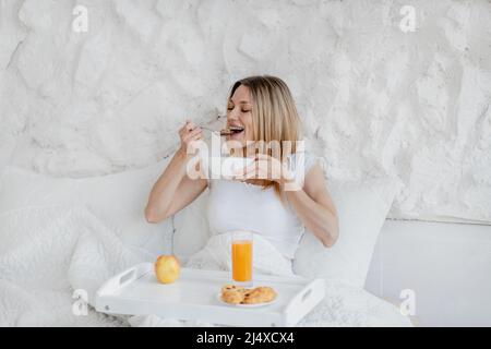 Felice abbastanza affamata giovane donna bionda europea svegliarsi, fare colazione in letto bianco Foto Stock