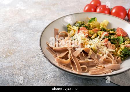 Piatto sano di tagliatelle a grani interi con verdure come broccoli, pomodori e spinaci, servito con parmigiano in un piatto su un rustico grigio Foto Stock