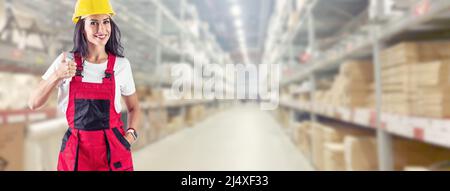 Un addetto al magazzino punta il dito verso l'alto. Sorridente giovane donna in abiti da lavoro di fronte ad un enorme magazzino. Foto Stock