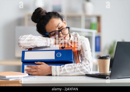 Donna d'affari stanca che dorme su una pila di cartelle alla sua scrivania di fronte al laptop, sentendosi esausta in ufficio Foto Stock